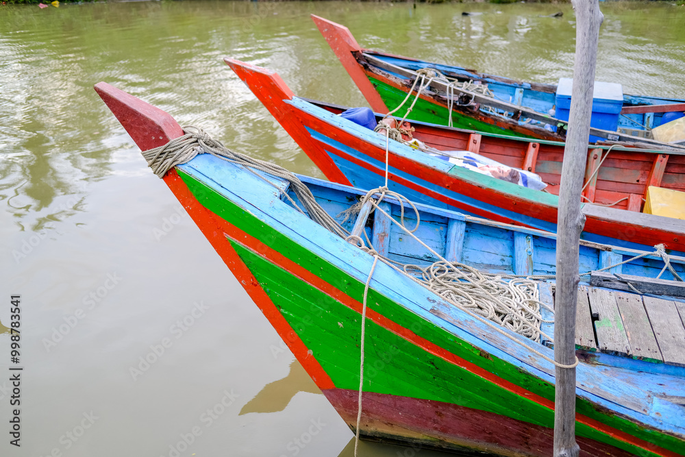 Colorful fishing boats docked along the concrete pier at Pelabuhan Kuala Singkawang, Kalimantan Barat, with village buildings and lush trees in the background, capturing coastal life.