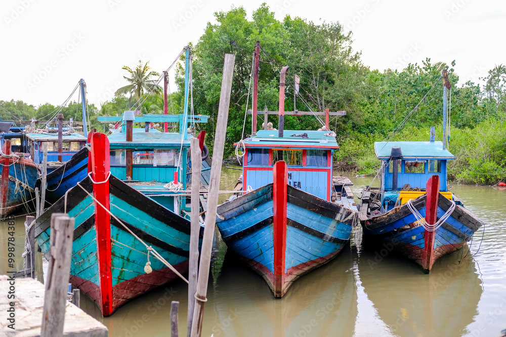 Colorful fishing boats docked along the concrete pier at Pelabuhan Kuala Singkawang, Kalimantan Barat, with village buildings and lush trees in the background, capturing coastal life.