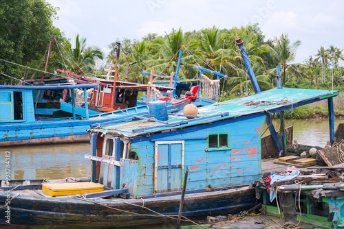 Obraz na płótnie Colorful fishing boats docked along the concrete pier at Pelabuhan Kuala Singkawang, Kalimantan Barat, with village buildings and lush trees in the background, capturing coastal life. w salonie