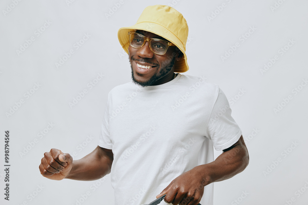 Smiling young man wearing a yellow bucket hat and glasses, casual white t shirt, posing joyfully against a light grey background, creating an upbeat and positive atmosphere Smiling young man wearing a yellow bucket hat and glasses, casual white t shirt, posing joyfully against a light grey background, creating an upbeat and positive atmosphere