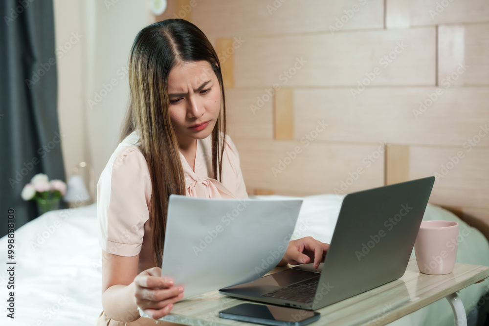 Asian woman reviewing document while working on laptop at home wearing pink blouse focused expression modern home office workspace freelance environment professional working from bedroom.