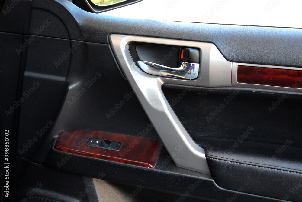 A close-up of a car door with black leather upholstery and wood trim. The combination of materials gives a rich, luxurious look to the car's exterior. Passenger door handle in cars interior.