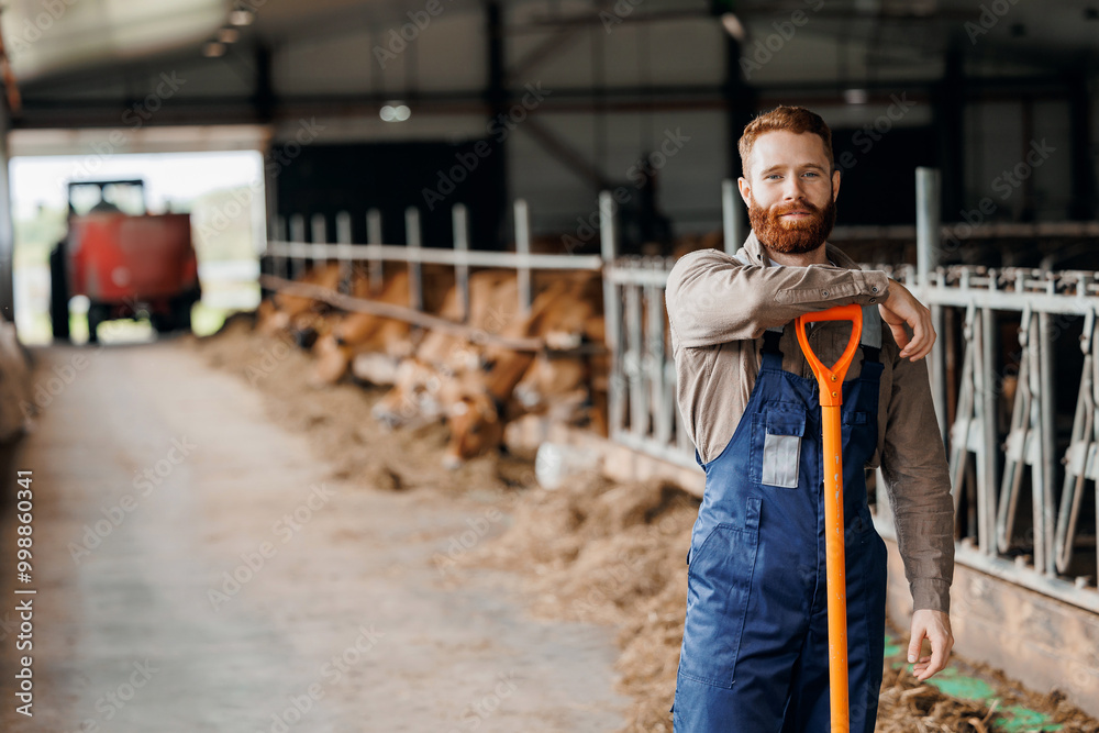 Portrait handsome young bearded farmer man in uniform against background of pen with cow. Concept worker of cattle livestock industry. Portrait handsome young bearded farmer man in uniform against background of pen with cow. Concept worker of cattle livestock industry.
