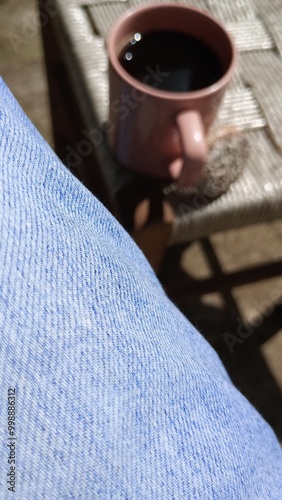 POV of a person sitting next to a coffee cup and a cookie. A man having an instant coffee cup. A cookie next to a coffee. A person wearing jeans. A coffee on a chair, under the sun. A coffee day.
