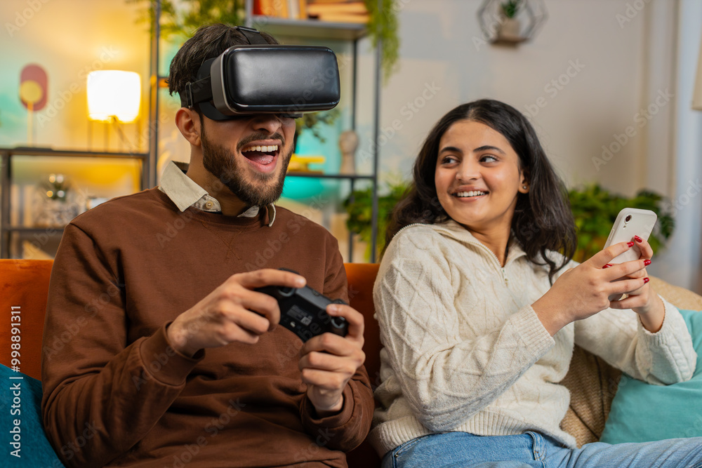 Happy amazed man wearing VR goggles playing videogame with joystick sitting beside Indian girlfriend using smartphone sitting on sofa in living room at home. Happy family spending weekend in apartment Happy amazed man wearing VR goggles playing videogame with joystick sitting beside Indian girlfriend using smartphone sitting on sofa in living room at home. Happy family spending weekend in apartment