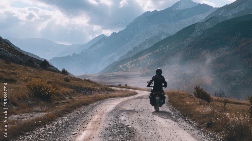 A lone motorcyclist rides down a winding dirt road through a vast mountainous landscape, bathed in misty light. A lone motorcyclist rides down a winding dirt road through a vast mountainous landscape, bathed in misty light.