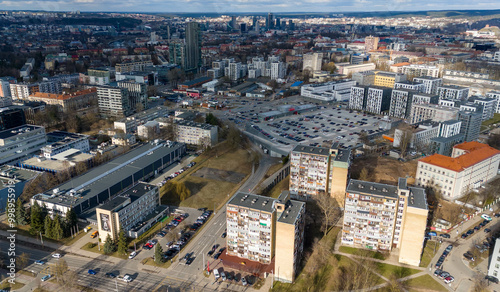 Aerial view of a modern urban area featuring a mix of residential and commercial buildings, with green spaces and parking lots. The skyline includes various structures.