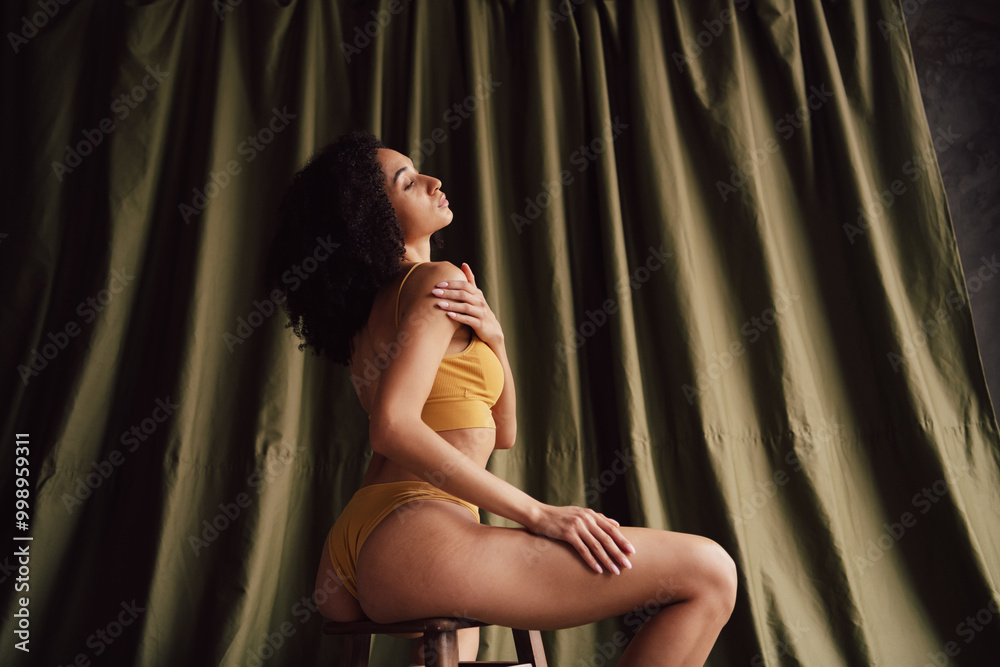 Unretouched portrait of gorgeous model girl sit chair touch shoulder khaki curtain background Unretouched portrait of gorgeous model girl sit chair touch shoulder khaki curtain background