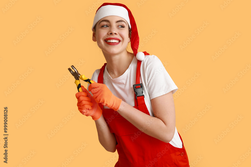Young female construction worker in Santa hat with pliers on yellow background Young female construction worker in Santa hat with pliers on yellow background