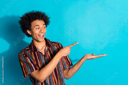 Photo of young funky guy with curly hair wearing summer hawaii shirt direct finger holding modern logo design isolated on blue color background photo