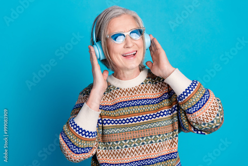 Photo of retired woman with white gray hairstyle wear ornament sweater in glasses touch headphones isolated on blue color background photo