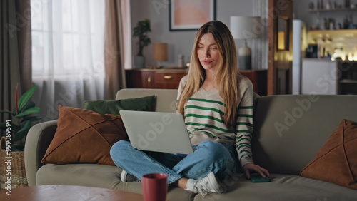 Remote worker reading message at smartphone sitting home couch with laptop.