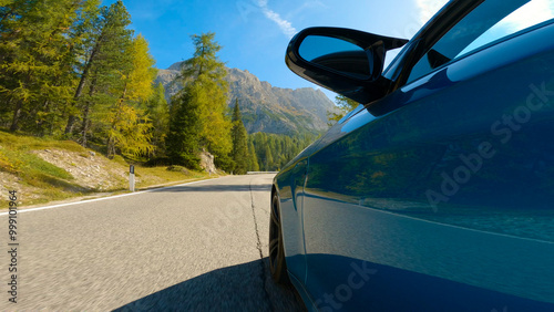 LOW ANGLE, CLOSE UP: Awesome wheel shot of a sportscar racing with a stunning view of the sunlit mountains of Italy. Blue car speeds along the breathtaking mountain road in the spectacular Dolomites. photo