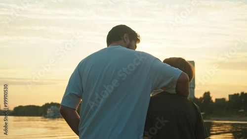 A young father provides support for his son. He embraces him, and together they observe the beautiful sunset over the lake.