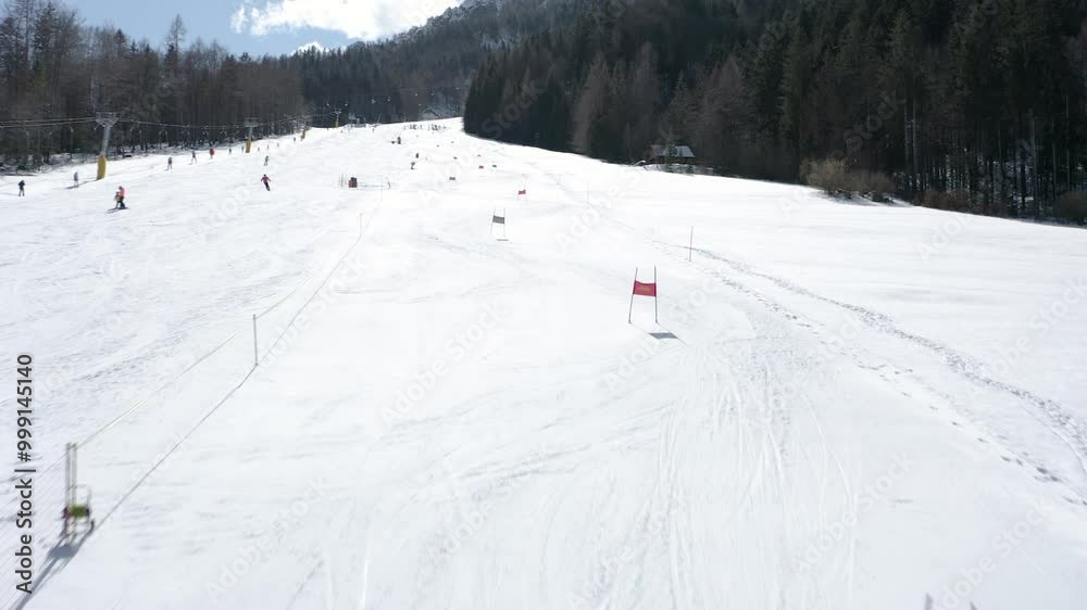 Ski slope under a clear sky with a racer navigating through the course marked by poles. Forested mountains line the background while other skiers make their way downhill. Ski slope under a clear sky with a racer navigating through the course marked by poles. Forested mountains line the background while other skiers make their way downhill.