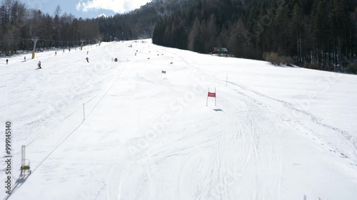 Ski slope under a clear sky with a racer navigating through the course marked by poles. Forested mountains line the background while other skiers make their way downhill.