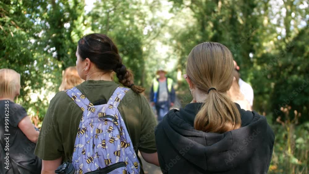 A group of people is walking along a tree-lined path, listening to a guide who is speaking ahead of them. The image captures an outdoor educational activity, such as a nature walk or guided tour A group of people is walking along a tree-lined path, listening to a guide who is speaking ahead of them. The image captures an outdoor educational activity, such as a nature walk or guided tour