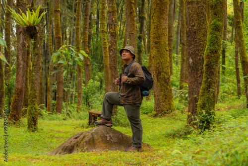 Asian man in adventure outfit in front of a big rock in a dense and green pine forest area. An adventurer in a flannel shirt, bucket hat, and backpack poses against a backdrop of dense pine trees.