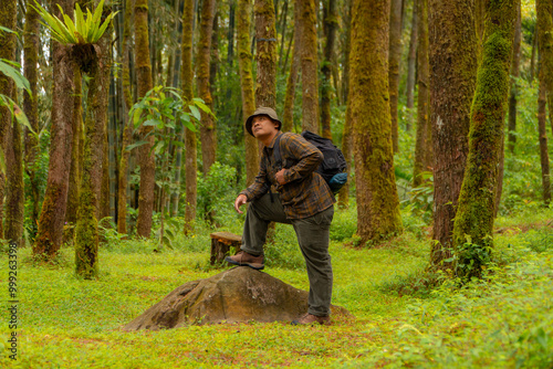 Asian man in adventure outfit in front of a big rock in a dense and green pine forest area. An adventurer in a flannel shirt, bucket hat, and backpack poses against a backdrop of dense pine trees.