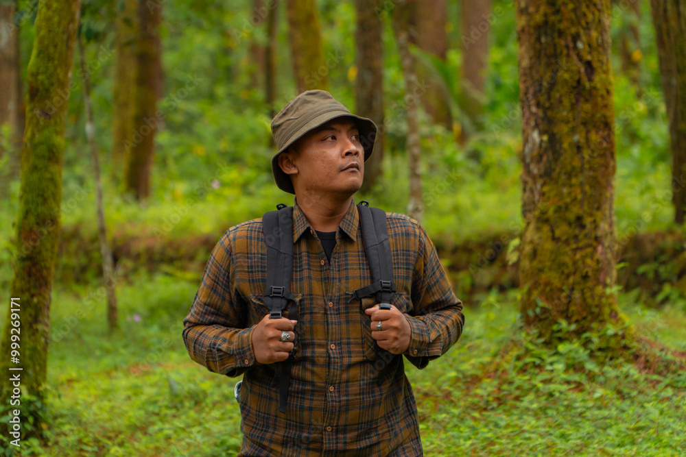 Half-body close-up of an adventurer in a dense and green pine forest. An Asian male traveler in a flannel shirt, bucket hat and backpack poses against a backdrop of dense pine trees. Half-body close-up of an adventurer in a dense and green pine forest. An Asian male traveler in a flannel shirt, bucket hat and backpack poses against a backdrop of dense pine trees.