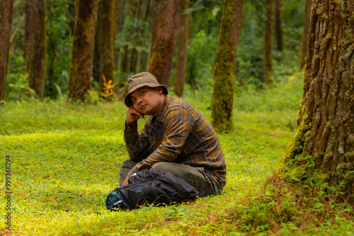 An adventurer is sitting on green grass in a lush pine forest area. Asian man in flannel shirt and bucket hat is sitting and resting while enjoying the tranquility of beautiful nature