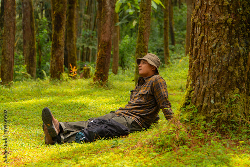 An adventurer is sitting on green grass in a lush pine forest area. Asian man in flannel shirt and bucket hat is sitting and resting while enjoying the tranquility of beautiful nature