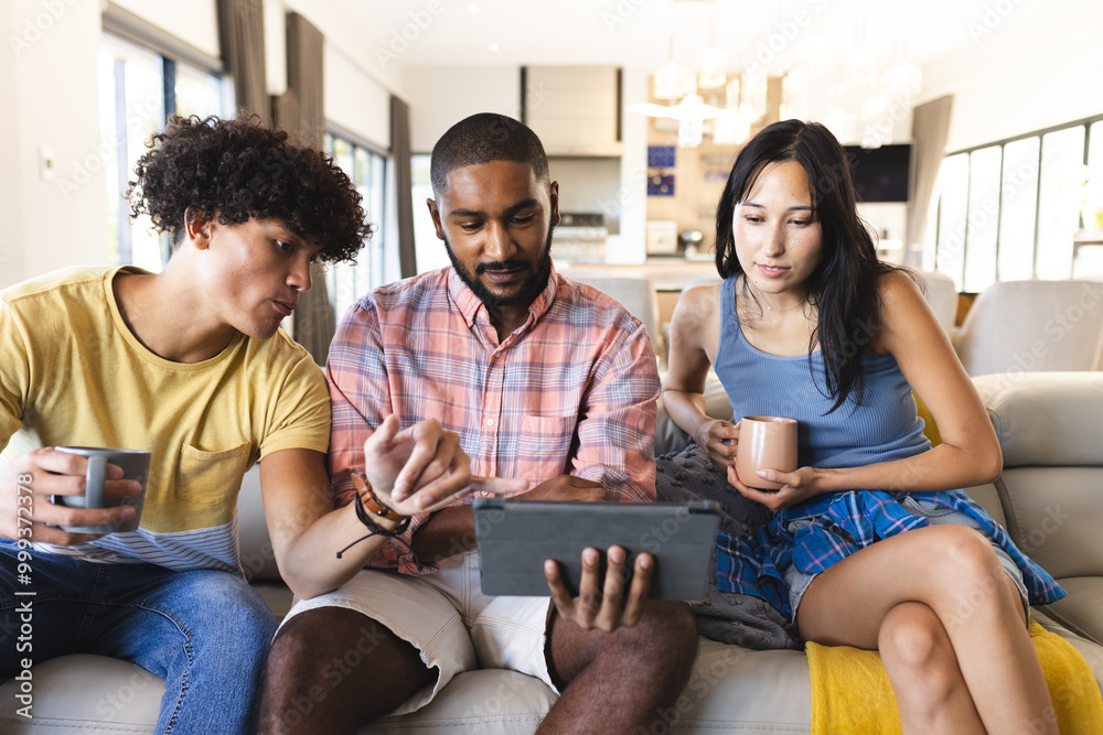Diverse friends hanging out, using tablet and drinking coffee on couch in living room, at home
