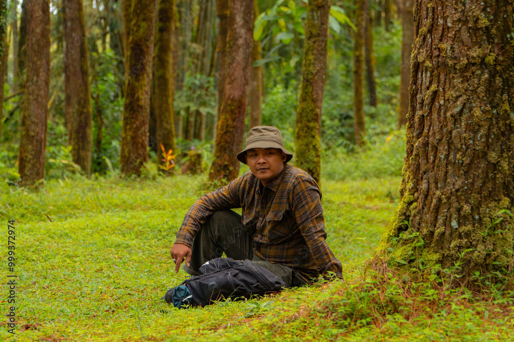 An adventurer is sitting on green grass in a lush pine forest area. Asian man in flannel shirt and bucket hat is sitting and resting while enjoying the tranquility of beautiful nature An adventurer is sitting on green grass in a lush pine forest area. Asian man in flannel shirt and bucket hat is sitting and resting while enjoying the tranquility of beautiful nature