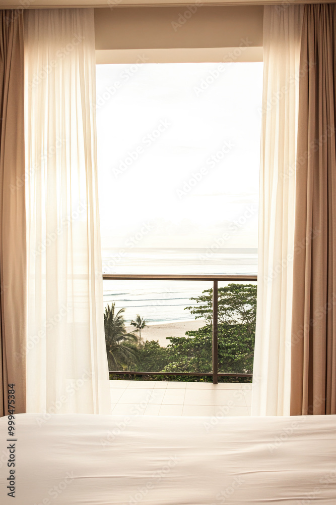 Fototapeta premium Coastal view through sheer curtains in a tranquil room with a balcony overlooking the ocean during early morning light