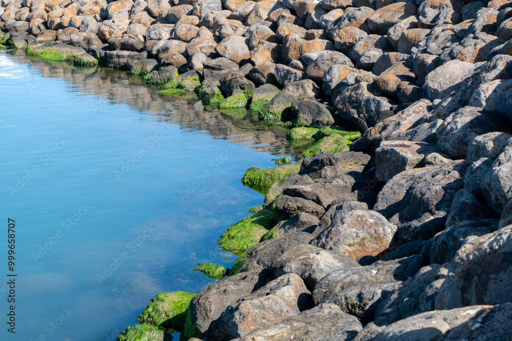 A rocky shoreline in Portarlington, Bellarine Peninsula of Victoria, Australia, with large boulders lining the water’s edge as part of a breakwater or sea wall to protect infrastructure from erosion. A rocky shoreline in Portarlington, Bellarine Peninsula of Victoria, Australia, with large boulders lining the water’s edge as part of a breakwater or sea wall to protect infrastructure from erosion.
