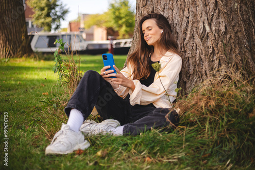 Portrait of smiling young brunette woman sitting on ground sitting under tree and reading text message on her cell phone outside. Laughing happy girl.