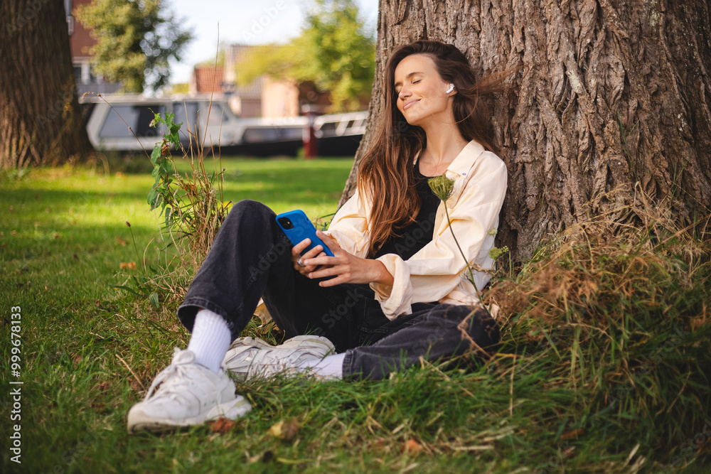 Woman sitting under tree in park, wear shirt, top, sneakers and jeans, hold mobile phone, earphones in ear. She turns on music, podcast or radio on her phone and listens to it with headphones. Woman sitting under tree in park, wear shirt, top, sneakers and jeans, hold mobile phone, earphones in ear. She turns on music, podcast or radio on her phone and listens to it with headphones.