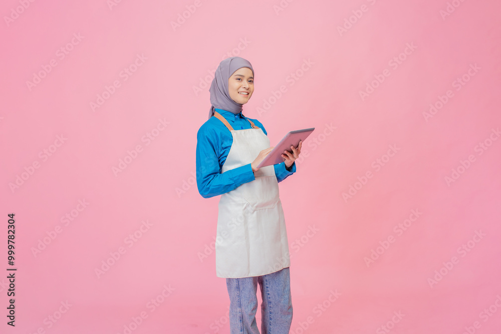 Fototapeta premium A joyful young woman in a stylish apron holds a tablet, smiling against a vibrant pink backdrop