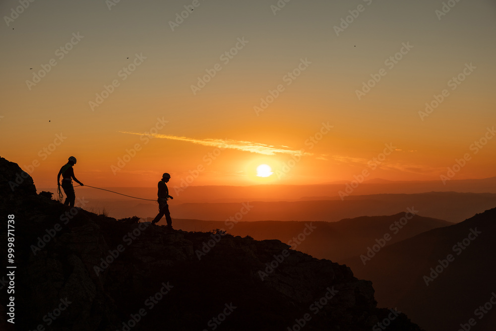 Two people are walking on a mountain top at sunset. The sky is orange and the sun is setting Two people are walking on a mountain top at sunset. The sky is orange and the sun is setting