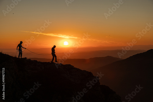 Two people are on a mountain, one of them is holding a rope. The sun is setting in the background, creating a beautiful and serene atmosphere