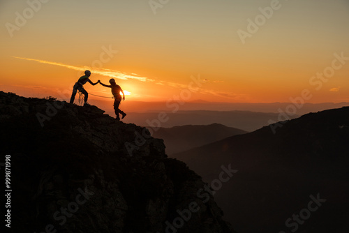 Two people are climbing a mountain together, one of them is helping the other. The sun is setting in the background, creating a beautiful and serene atmosphere
