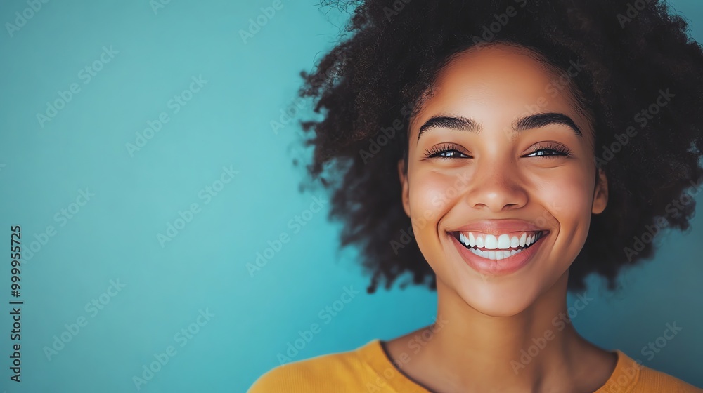 A young woman with curly black hair smiles brightly at the camera against a blue background. A young woman with curly black hair smiles brightly at the camera against a blue background.