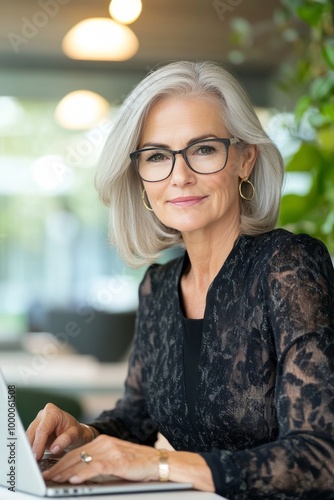 A professional woman with gray hair works on a laptop in a modern office fill...