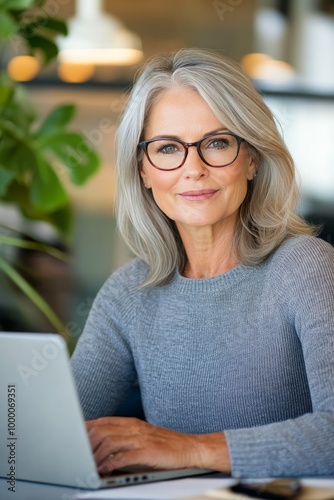 A professional woman with gray hair works on a laptop in a modern office fill...