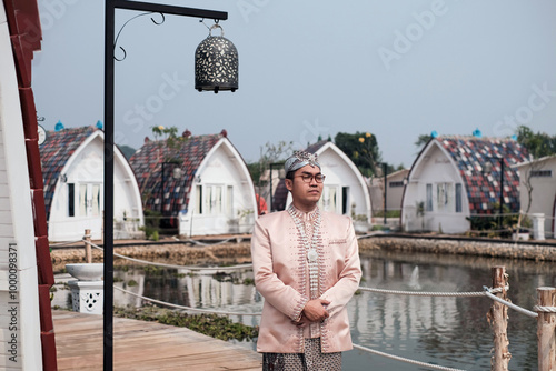 The groom wearing a light brown beskap adorned with elegant decorations, perfect for a traditional Indonesian wedding from West Java, Sunda, with a serene lake and traditional rice barn (lumbung padi) photo