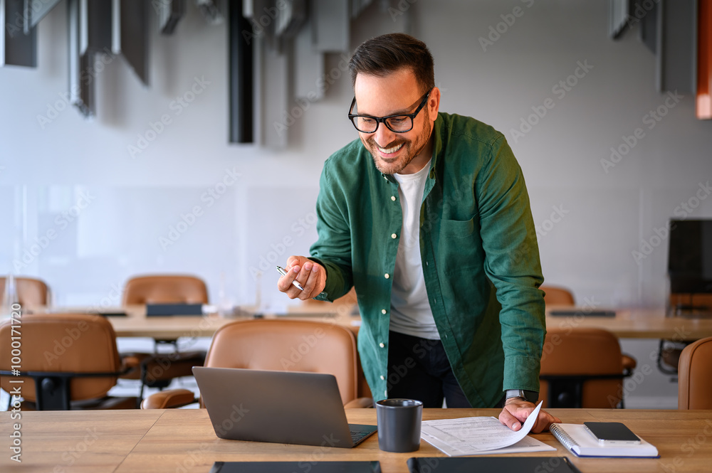 Positive businessman smiling and discussing over video call on laptop while standing at desk in office Positive businessman smiling and discussing over video call on laptop while standing at desk in office