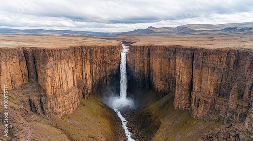 A narrow waterfall drops dramatically into a steep canyon, surrounded by rugged cliffs and dense wilderness under a cloudy sky Waterfall plunge wilderness, Wild canyon serenity