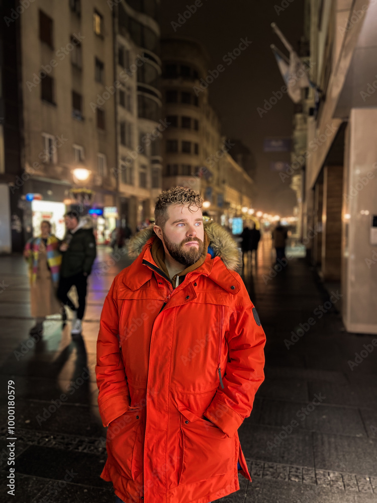 Fototapeta premium Man walking in a bustling nighttime city street, wearing a bright orange parka. Serene contemplation against the backdrop of lively city lights at night. Urban life concept