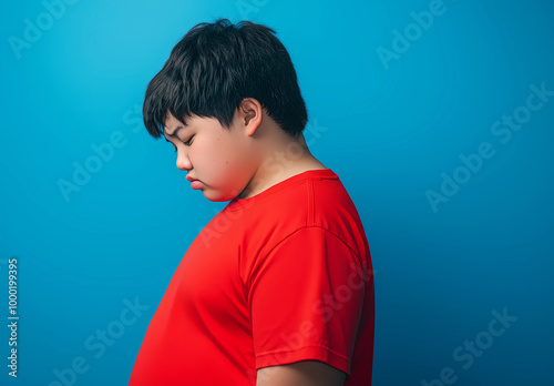 A side profile photograph of an overweight teen boy wearing a red t-shirt, standing with his head down, looking sad and dejected against a blue background photo