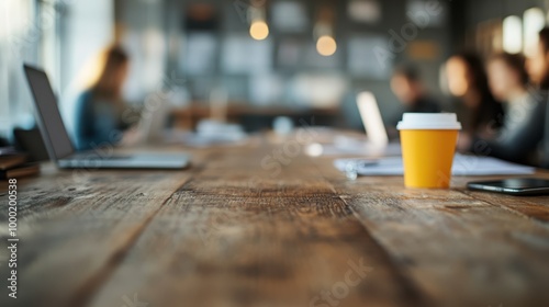 A cozy office setting featuring a wooden table, orange coffee cup, and blurred background of employees working at their laptops, conveying a productive workspace.