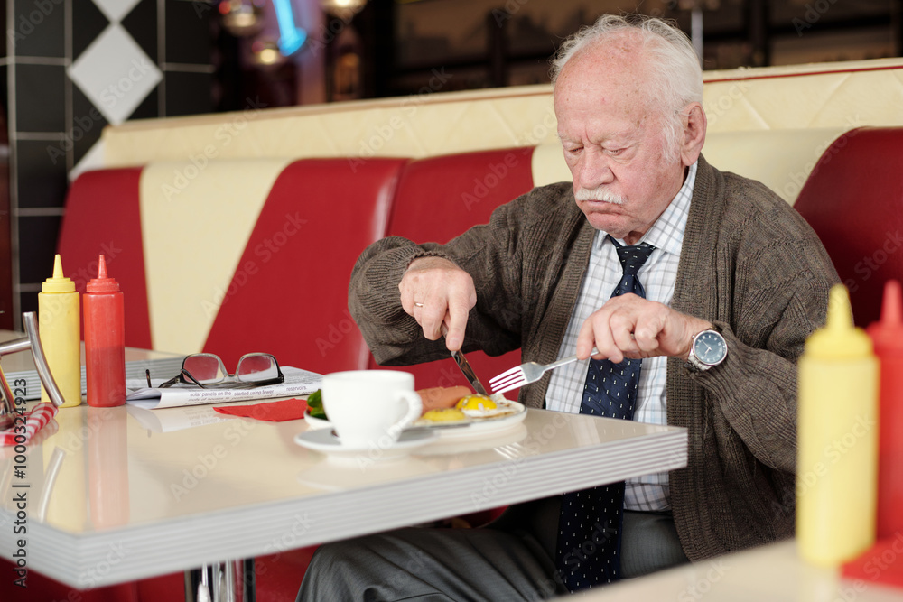 Elderly man enjoying meal in retro-style diner, with red and white seating accents and condiments on table. He is wearing a tie and sweater while focusing on his food