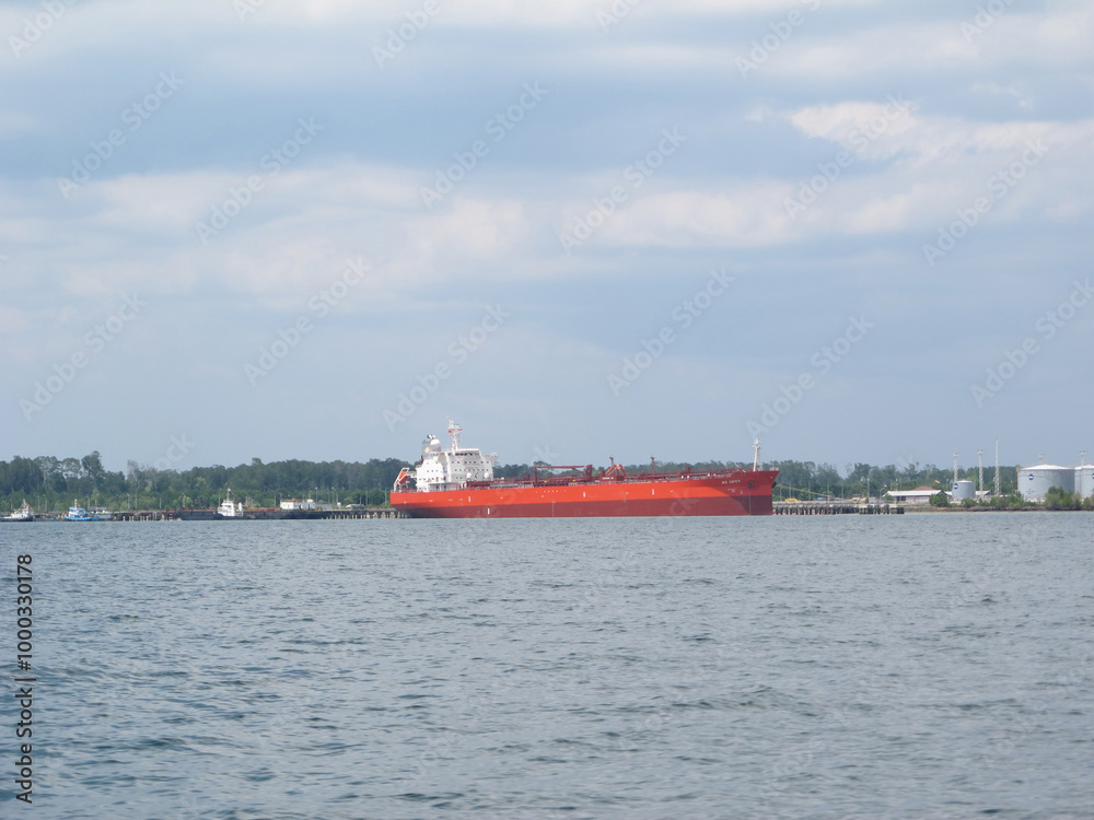 Fototapeta premium A large industrial cargo ship anchored in calm waters under a bright blue sky. The ship, with its distinct red and black hull, is equipped with towering cranes and communication masts
