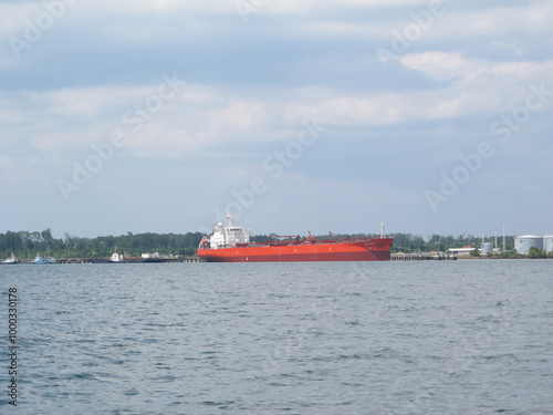 A large industrial cargo ship anchored in calm waters under a bright blue sky. The ship, with its distinct red and black hull, is equipped with towering cranes and communication masts