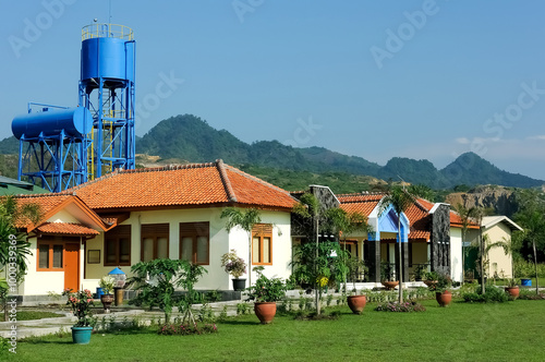 A serene villa surrounded by lush greenery, featuring a traditional yet modern architectural design with a sloping red-tiled roof. The villa is nestled amidst a garden landscape