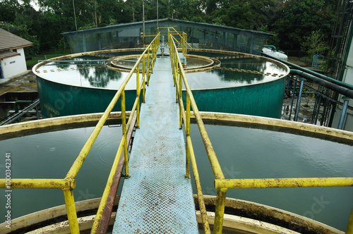 A Water Treatment with a walkway bridge leading to a series of large, circular water treatment tanks at an industrial facility. The yellow handrails on either side of the blue metallic walkway provide photo